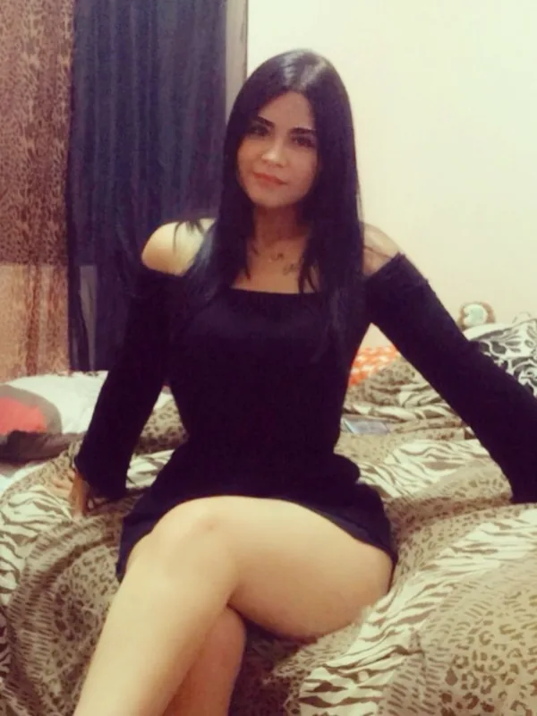Femme aux cheveux noirs portant une robe noire, assise sur un lit à motifs animaliers, regardant l'objectif.